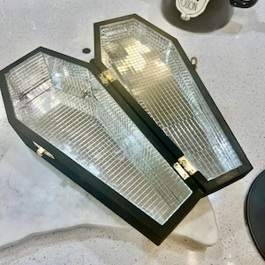 May include: A black wooden coffin-shaped box with a mirrored interior. The box is open and the lid is propped up. The box has a gold latch and hinges.