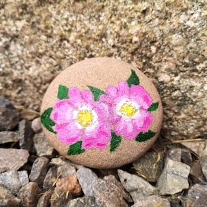May include: A hand-painted, round stone featuring two pink flowers with yellow centers and green leaves. The stone is a light brown color and rests on a bed of small rocks. The flowers are the focal point of this nature-inspired art piece.