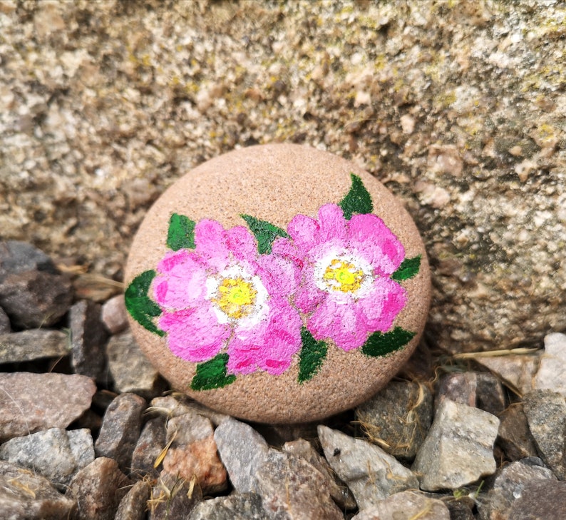 May include: A hand-painted, round stone featuring two pink flowers with yellow centers and green leaves. The stone is a light brown color and rests on a bed of small rocks. The flowers are the focal point of this nature-inspired art piece.