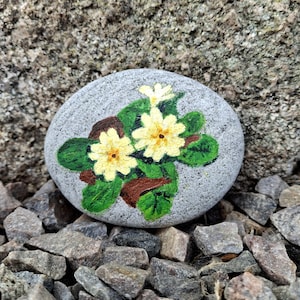 May include: A hand-painted, gray stone featuring a floral design. The artwork depicts two yellow flowers with brown centers and green leaves. The stone is surrounded by small rocks, creating a natural, decorative piece.