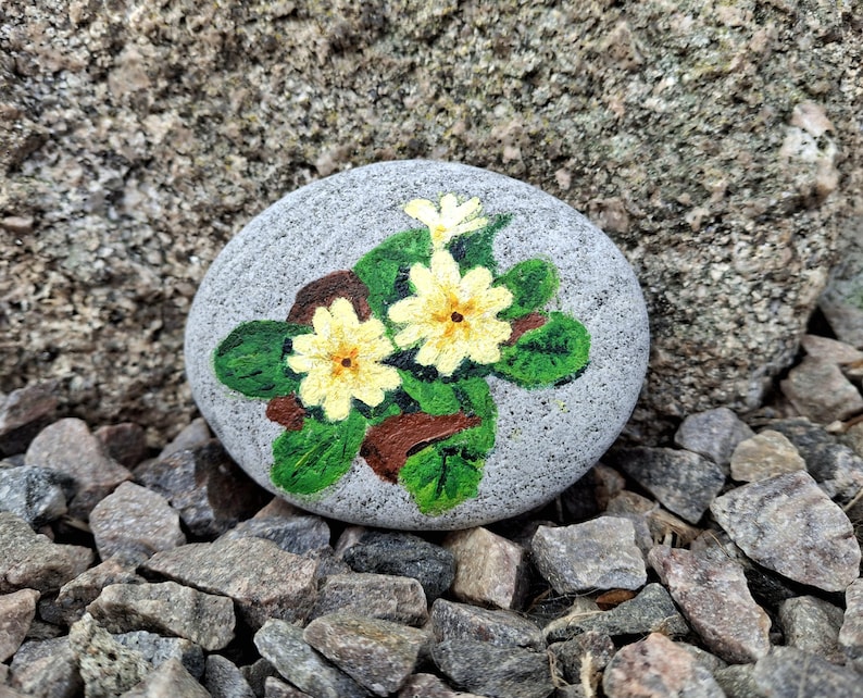 May include: A hand-painted, gray stone featuring a floral design. The artwork depicts two yellow flowers with brown centers and green leaves. The stone is surrounded by small rocks, creating a natural, decorative piece.