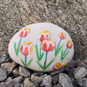 May include: A painted stone featuring a floral design of red and yellow tulips with green stems and leaves. The artwork is on a smooth, light gray stone, resting on a bed of small rocks, with a larger stone in the background.
