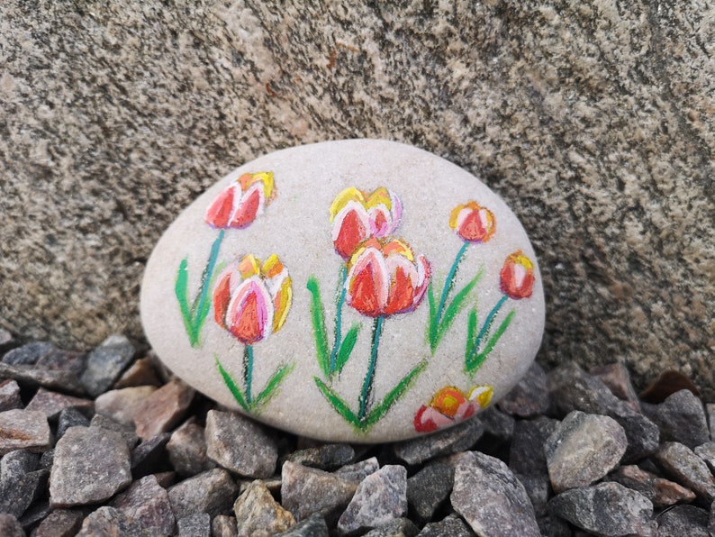 May include: A painted stone featuring a floral design of red and yellow tulips with green stems and leaves. The artwork is on a smooth, light gray stone, resting on a bed of small rocks, with a larger stone in the background.