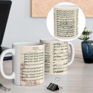 May include: Two white ceramic mugs with a floral design and musical notes printed on them. The mugs are on a wooden table with a laptop, a binder clip, and a potted plant in the background.