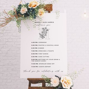 May include: A white wedding welcome sign with black text and a floral monogram. The sign lists the wedding day schedule, including the ceremony, photos, dinner, speeches, first dance, cake cutting, and the send-off. The text reads "HANAH + ALEX HARRIS welcome you".