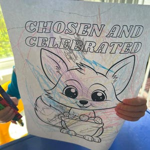 Op de afbeelding: Een kleurplaat met de tekst "CHOSEN AND CELEBRATED" boven een cartoon vos illustratie. De vos is gekleurd met verschillende tinten, en de pagina heeft kleurpotloodstrepen. De afbeelding wordt door een persoon omhoog gehouden.