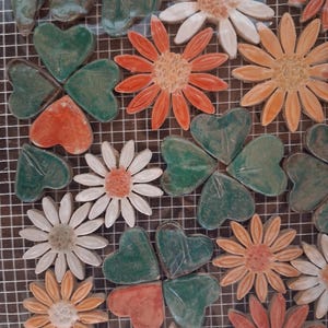 Puede incluir: Una colección de azulejos de mosaico de cerámica dispuestos sobre una rejilla blanca. Las piezas incluyen flores en forma de margarita blancas, naranjas y color melocotón con centros texturizados, junto a hojas en forma de corazón.
