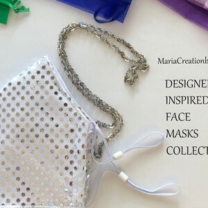 May include: A white face mask with silver sequins and a silver chain. The mask has adjustable ear loops and is part of the "DESIGNERS INSPIRED FACE MASKS COLLECTION" by MariaCreationbyMaria.