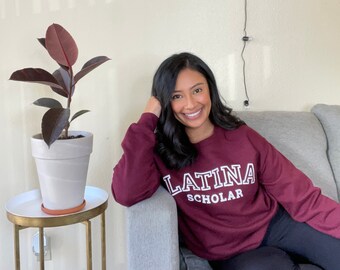 Op de afbeelding: Een vrouw draagt een bordeauxrood sweatshirt met de tekst "LATINA SCHOLAR" in witte letters. Ze zit op een grijze bank met een kamerplant in een witte pot links.