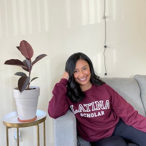 May include: A woman wearing a maroon sweatshirt with the text "LATINA SCHOLAR" in white lettering. She is sitting on a gray couch with a houseplant in a white pot to the left.