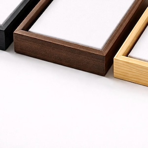 Puede incluir: Primer plano de tres marcos rectangulares de madera con inserciones de lienzo blanco. Los marcos son de diferentes tonos de madera: marrón oscuro, marrón claro y negro. Los marcos están dispuestos uno al lado del otro sobre un fondo blanco.