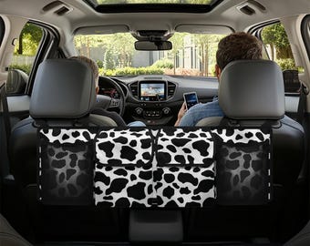 May include: Black and white cow print car seat back organizer with multiple pockets. The organizer is hanging from the headrests of the back seats of a car.