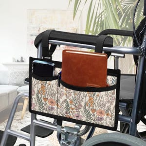 May include: Wheelchair side bag with a floral pattern in cream, orange, and green. The bag has multiple pockets, holding a brown notebook, a smartphone, and a pen. The bag is attached to the armrest of a wheelchair.