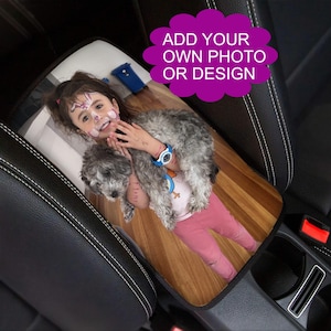 May include: Custom car seat cover featuring a photo of a child with a dog. The cover has a wooden floor design and the text "ADD YOUR OWN PHOTO OR DESIGN" in a purple cloud shape. The cover is placed on a black car seat.