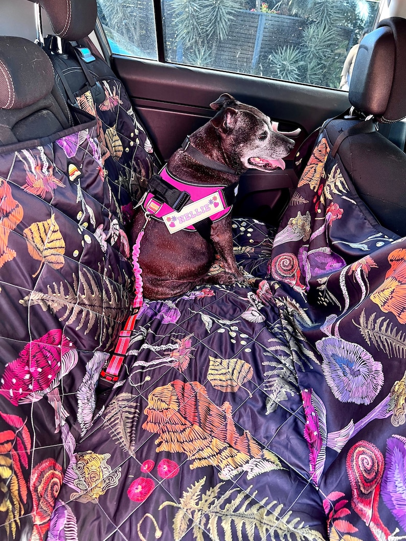 May include: A black and pink dog harness with the name "Nellie" printed on it. The harness is on a dog sitting in the back seat of a car. The car seat is covered with a black quilted fabric with a colorful floral pattern.