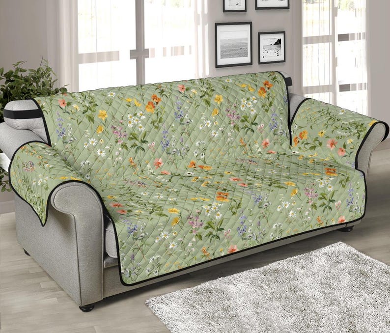 May include: A green quilted sofa cover with a floral pattern featuring a variety of flowers in shades of pink, yellow, and white. The cover has a black trim and is designed to protect furniture from spills and stains.