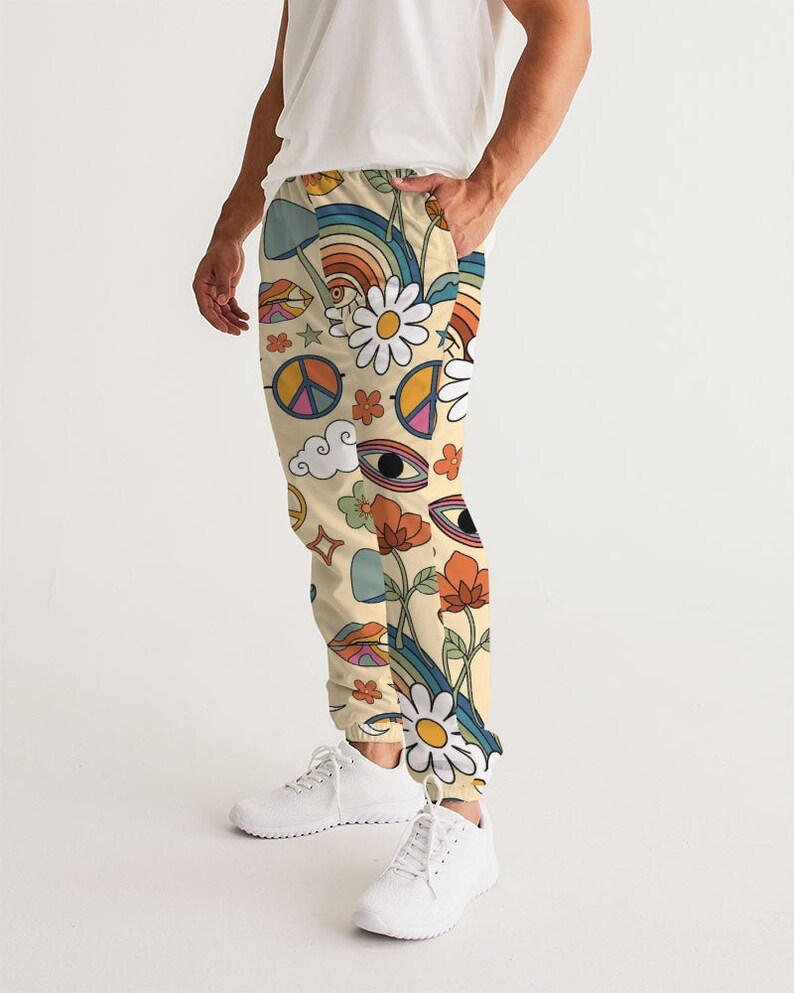 Puede incluir: Un pantal&oacute;n de ch&aacute;ndal de color crema con un estampado retro colorido que incluye s&iacute;mbolos de paz, flores, arco&iacute;ris y ojos.