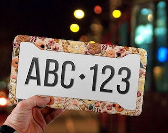 May include: A rectangular license plate frame featuring a beige background with a dense, colorful floral pattern in shades of orange, pink, yellow, and green. The center of the frame displays the black text ABC 123 with a diamond separator.