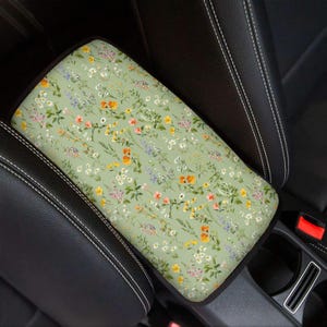 Può includere: Un copribracciolo per auto con motivo floreale. La copertura presenta uno sfondo verde chiaro ornato da un motivo di fiori selvatici colorati nei toni del giallo, arancione, rosa e viola. La copertura rettangolare è progettata per il bracciolo centrale dell'auto.