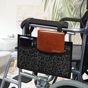 May include: A black and gray leopard print wheelchair side bag with multiple pockets. The bag is attached to the armrest and holds a brown notebook, a smartphone, and pens. The bag is designed for convenience and accessibility.