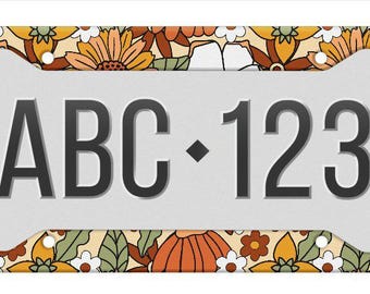 May include: A rectangular license plate frame featuring a repeating pattern of orange, yellow, and white flowers with green leaves on a cream background. The center of the frame is light gray and displays the black text ABC 123 with a diamond symbol.