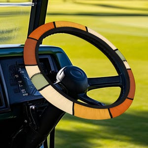 Könnte beinhalten: Ein farbenfroher Lenkradbezug mit einem geometrischen Muster in Orange-, Braun-, Grün- und Cremetönen. Der Bezug befindet sich auf einem schwarzen Lenkrad, vor einem grünen Hintergrund, wahrscheinlich einem Golfwagen.