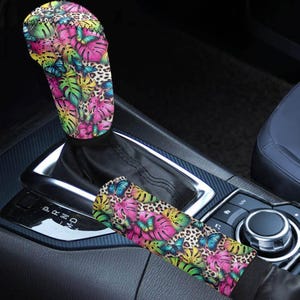 May include: A car gear shift cover and armrest cover set. The covers feature a vibrant pattern of pink, yellow, green, and blue butterflies and tropical leaves on a leopard print background. The covers are shown in a car interior.