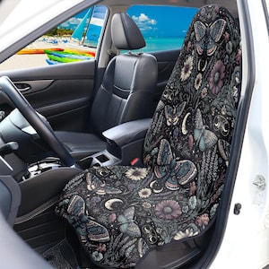 May include: Black car seat cover featuring a floral and butterfly design. The cover has a dark background with illustrations of butterflies, flowers, and crescent moons in shades of blue, purple, and white. The car seat cover is shown inside a car.