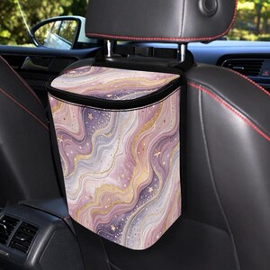 May include: A car trash bag with a black zipper and strap, featuring a swirling pattern in shades of purple, pink, and gold. The design includes small star accents. The bag is hanging from a car headrest.