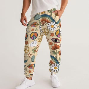 May include: Cream-colored joggers featuring a vibrant, repeating pattern of colorful rainbows, white daisies, orange flowers, peace signs, stylized eyes, lips, clouds, and stars. The pants have an elastic waistband and cuffed ankles.