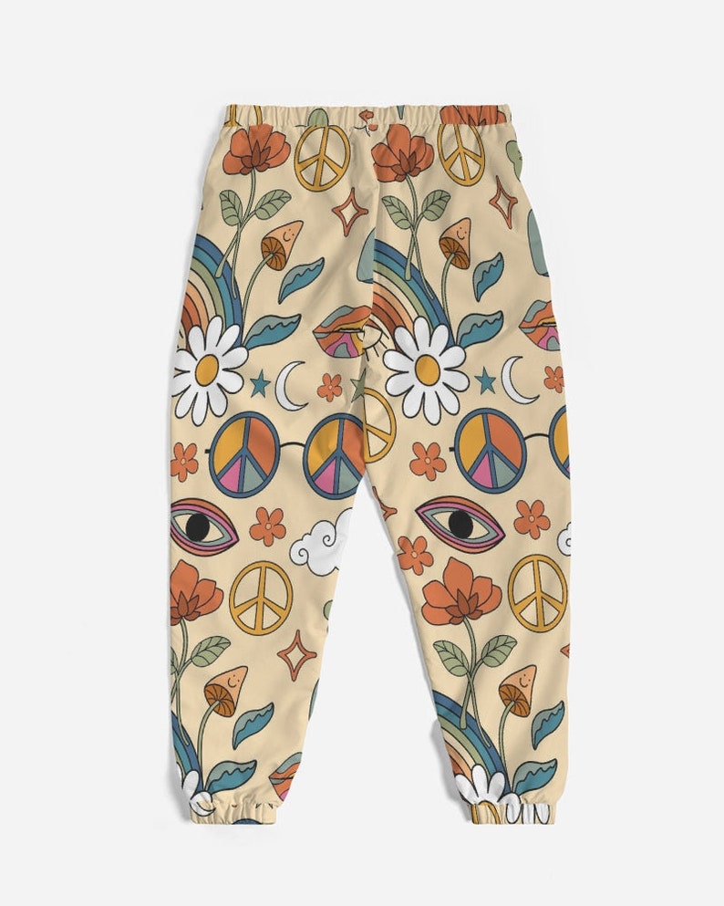 Puede incluir: Pantalones de ch&aacute;ndal beige con un estampado retro de los a&ntilde;os 70 con signos de paz, flores, arco&iacute;ris y ojos.