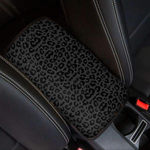 May include: A black and grey leopard print car armrest cover. The cover is rectangular and fits over the centre console armrest. The cover is surrounded by black leather car seats with white stitching.