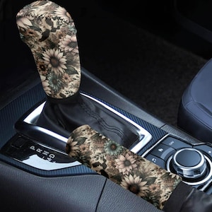 May include: A car gear shift cover and seat belt cover set. The covers feature a floral pattern with sunflowers, daisies, and other flowers in shades of beige, brown, and green. The covers are set against a black interior.