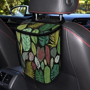 Op de afbeelding: Een zwarte auto afvalzak met een ritssluiting, hangend aan een hoofdsteun van een auto. De tas heeft een patroon van groene, gele en rode bladeren op een zwarte achtergrond. De tas is ontworpen om afval in te bewaren en de auto schoon te houden.