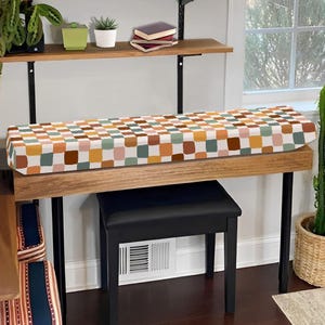 May include: A rectangular piano cover featuring a repeating pattern of rounded squares in shades of mustard yellow, sage green, terracotta orange, light pink, and white, draped over a wooden piano keyboard stand with a black bench underneath.