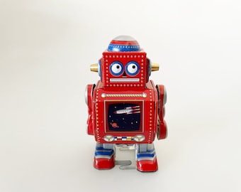 Peut inclure: Un robot jouet rouge à remonter avec des accents bleus et blancs. Le robot a une grosse tête avec des yeux globuleux et une petite antenne. Le corps du robot a une forme rectangulaire avec une fenêtre montrant une scène spatiale avec une fusée et des planètes. Le robot a des bras et des jambes qui bougent lorsqu'il est remonté.