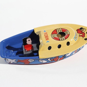 Op de afbeelding: Een geel en blauw speelgoedbootje met een rode reddingsboei en het woord "ROBIN" erop gedrukt. Het bootje heeft een klein figuurtje van een persoon aan de voorkant en een rode krab aan de zijkant.