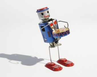 May include: A blue and red tin robot toy with a red hat and a drum. The robot has a smiling face and is holding a drum with red, yellow, and blue stripes.