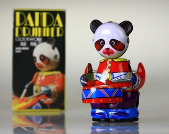 May include: A wind-up toy panda drummer with a red, white, and blue uniform, holding drumsticks and a drum with a red, blue, and yellow design. The toy is on a white surface with a box labeled "Panda Drummer" in the background.