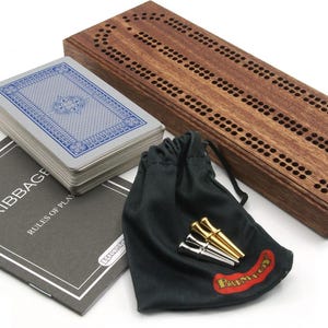 Op de afbeelding: Een houten cribbage-bord met een donkerbruine afwerking. Het bord heeft een baan om punten te scoren en een klein zakje voor pionnen. De set bevat ook een kaartspel en een boekje met regels.