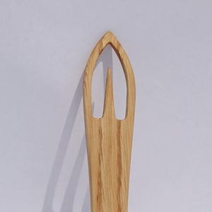 May include: A wooden knitting needle with a pointed tip and two loops at the bottom. The needle is made of light brown wood with a grain pattern.