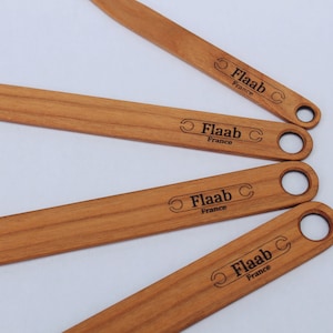 May include: Several wooden spatulas with a light brown finish are arranged on a white surface. Each spatula has a rounded handle with a hole and the text "Flaab France" printed on the front.