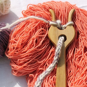 May include: A wooden knitting tool with two prongs, used to create a slip knot. The tool is surrounded by balls of yarn in white, purple, and orange.