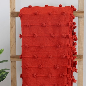 Könnte beinhalten: Eine leuchtend rote Decke mit strukturiertem Design und dekorativen Quasten. Die Decke hat horizontale Reihen mit erhabenen, geknoteten Details und Fransen an den Kanten. Sie ist über eine Holzleiter drapiert.