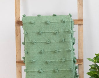 May include: A sage green throw blanket with a textured design and fringe detail. The blanket is draped over a wooden ladder. The blanket features rows of raised, knotted embellishments.