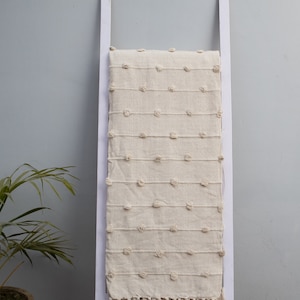 Puede incluir: Manta rectangular de color crema con un patrón punteado texturizado y detalles de flecos. La manta se muestra en una escalera blanca sobre un fondo gris. La manta está hecha de un material suave y natural.