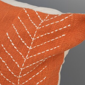 May include: Close-up of a decorative pillow with an orange cover and white stitching. The stitching forms a leaf-like pattern on the pillow's surface. The pillow has a white border.
