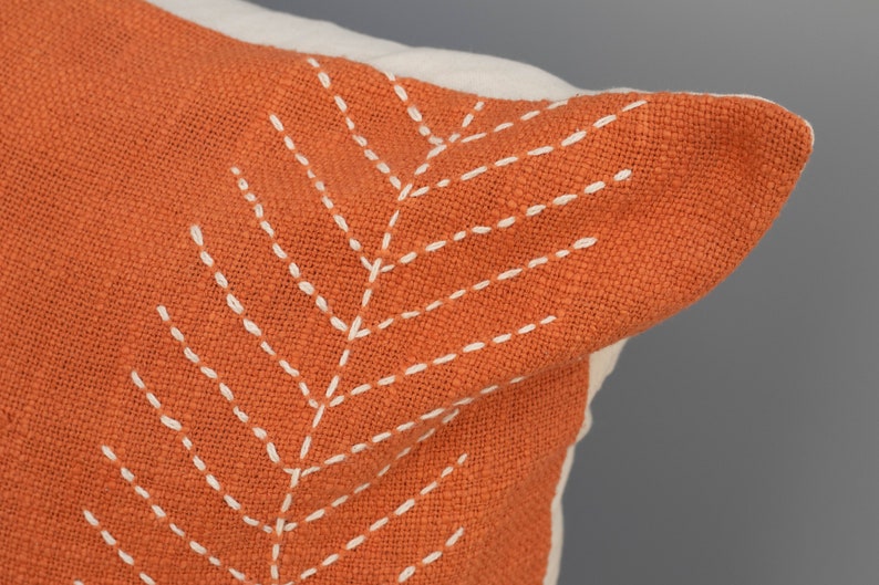 May include: Close-up of a decorative pillow with an orange cover and white stitching. The stitching forms a leaf-like pattern on the pillow's surface. The pillow has a white border.