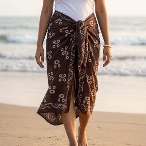 May include: A brown sarong with a white floral pattern tied at the waist. The sarong is worn over a white tank top. The person is walking on a sandy beach near the ocean.