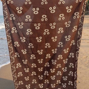 May include: A brown sarong with a white floral pattern and decorative border is held up on a beach. The sarong's design features clusters of white flowers evenly spaced across the fabric. The border has a white, swirling design.